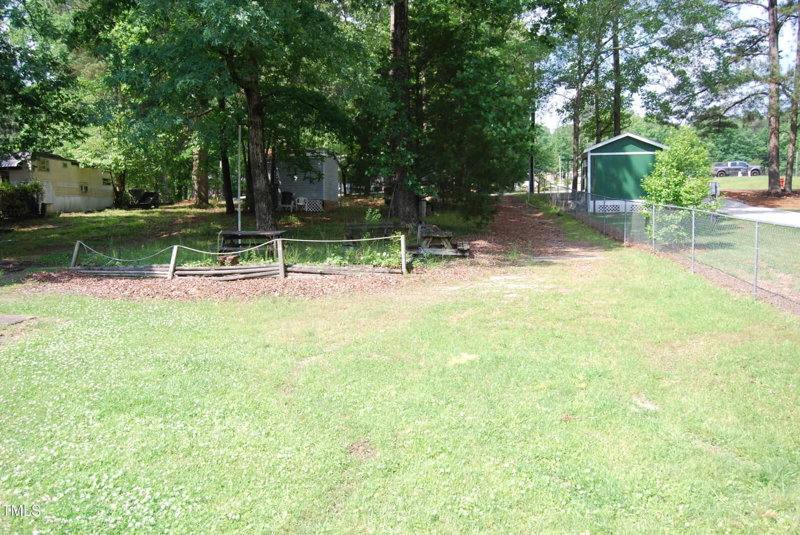 A grassy backyard with trees, a wooden garden bed, picnic tables, a shed, a wire fence, and a paved road visible on the right. Sunlight and shadows are present throughout the area.