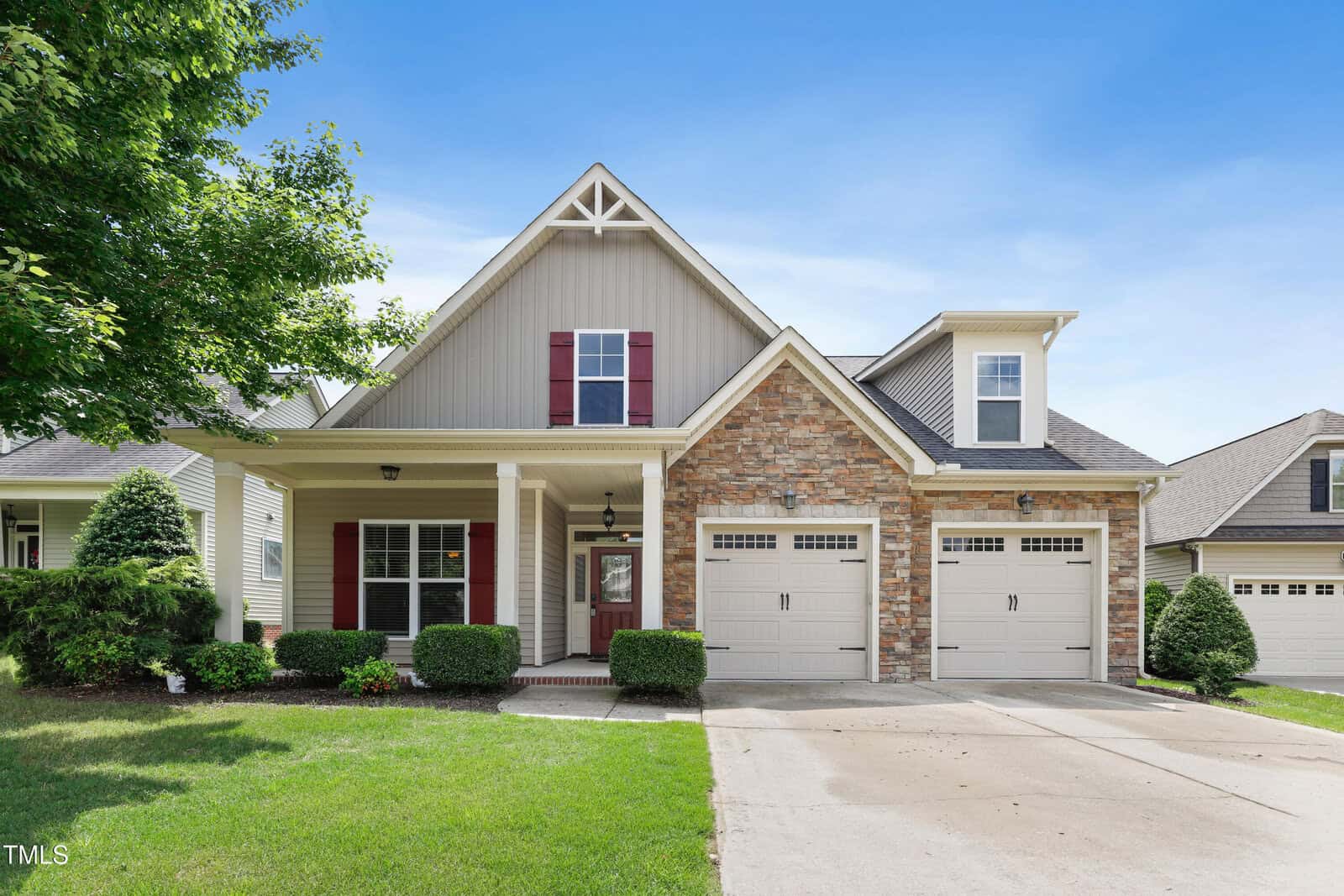 Single-family suburban house with a gable roof, stone and siding exterior, red shutters, two-car garage, front porch, and landscaped lawn under a blue sky.