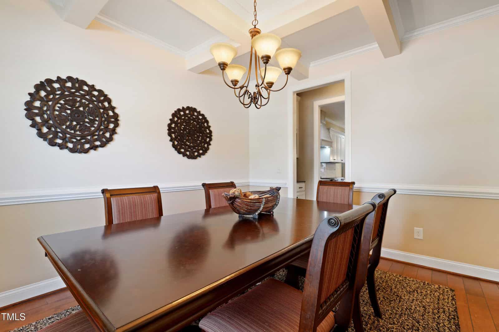 A dining room with a dark wooden table, six matching chairs, a decorative bowl centerpiece, two metal wall art pieces, a chandelier overhead, and neutral-colored walls with white trim.
