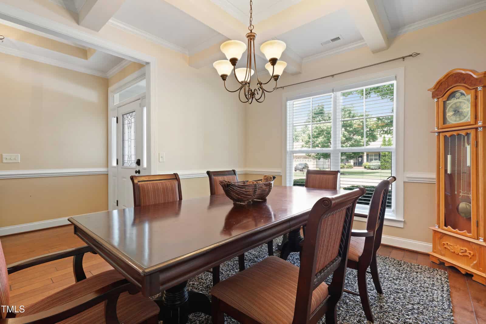 A dining room with a dark wood table and six chairs, a woven basket centerpiece, a window with blinds letting in daylight, a grandfather clock, a rug, and a chandelier hanging from a coffered ceiling.