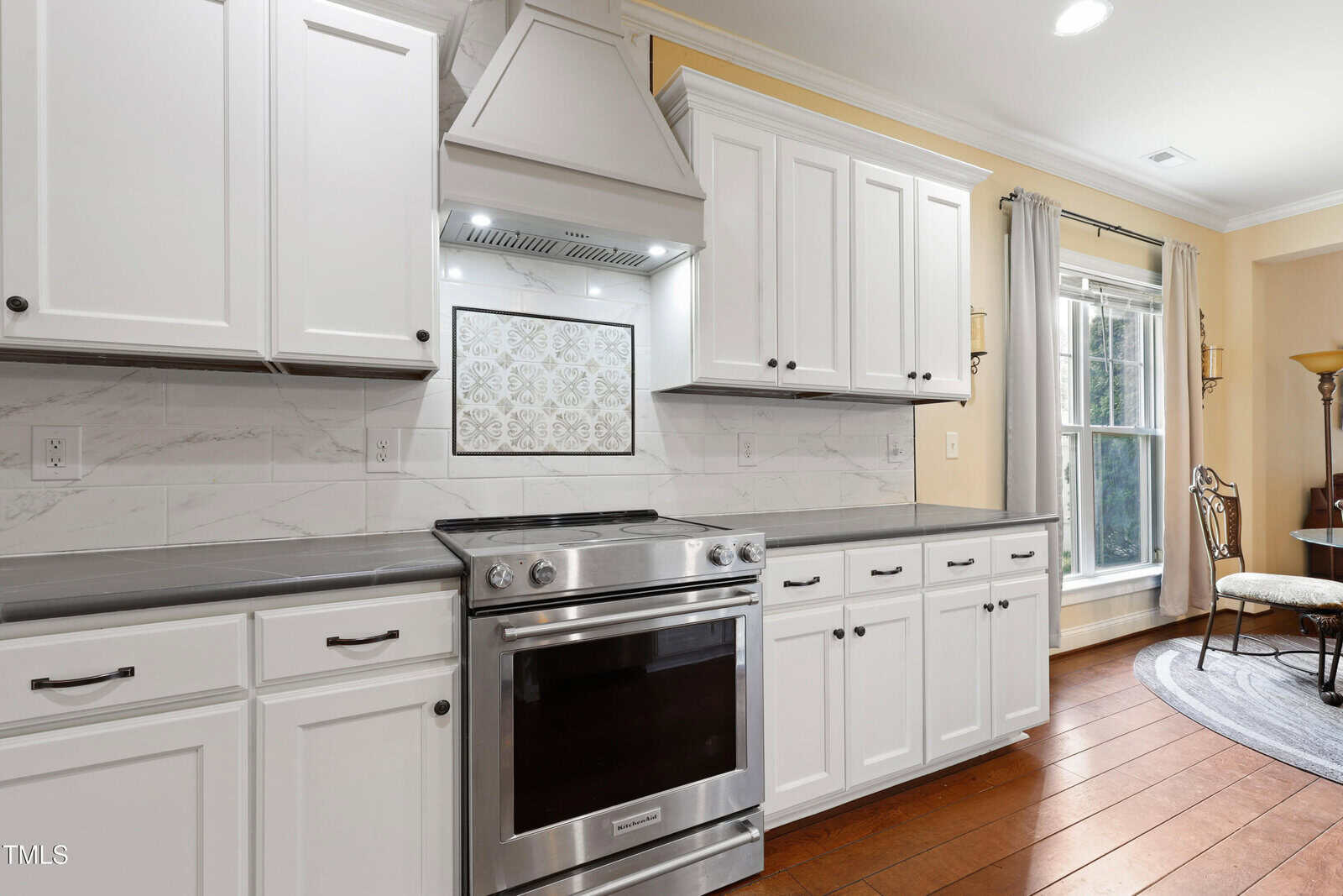 Modern kitchen with white cabinets, stainless steel oven and range hood, gray countertops, and decorative tile backsplash. Large window lets in natural light, and a dining area with a round table is visible to the right.