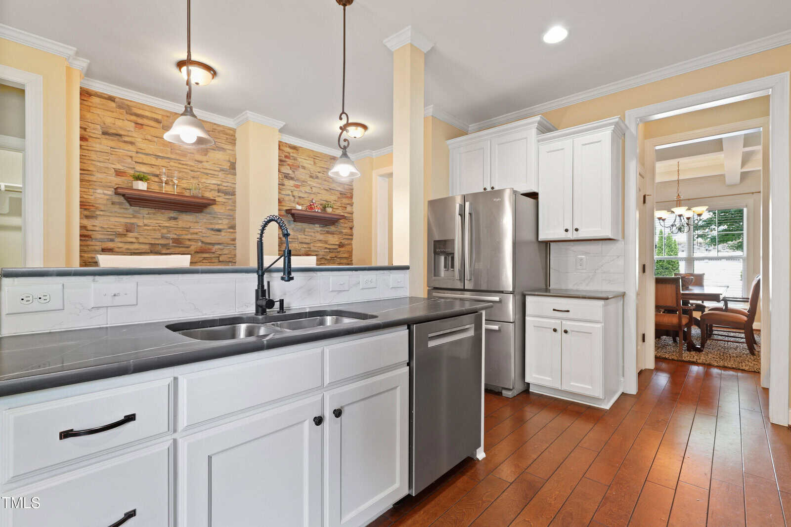 Modern kitchen with white cabinets, stainless steel appliances, black countertops, double sink, pendant lighting, and hardwood floors. A stone accent wall and dining area are visible in the background.