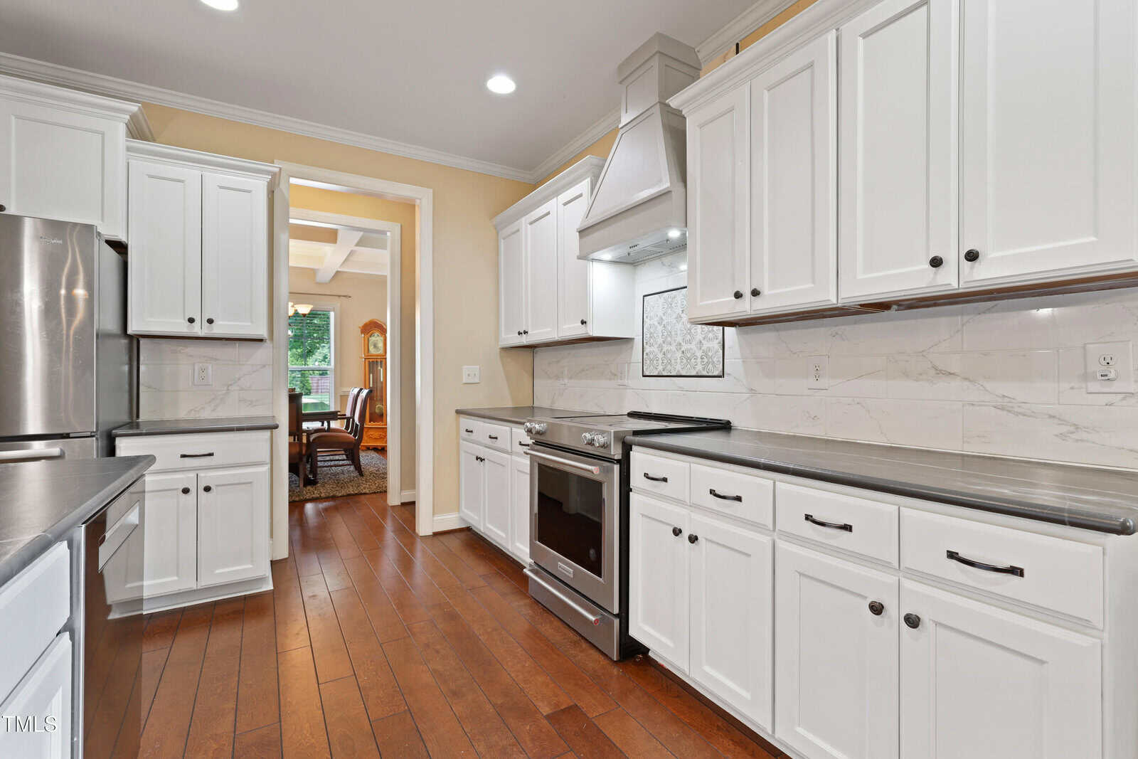 A modern kitchen with white cabinets, stainless steel appliances, dark countertops, wood floors, and marble backsplash. A dining area is visible through a doorway in the background.