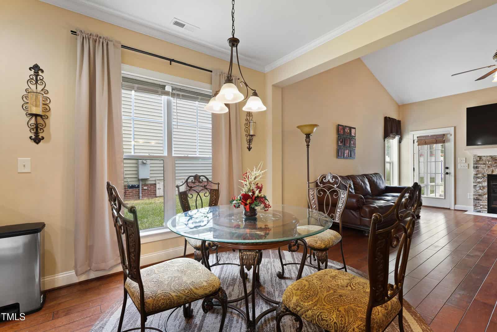 A dining area with a round glass table, four ornate chairs, a floral centerpiece, and a three-light chandelier. A window with beige curtains is on the left, and a living area with a leather sofa is visible in the background.