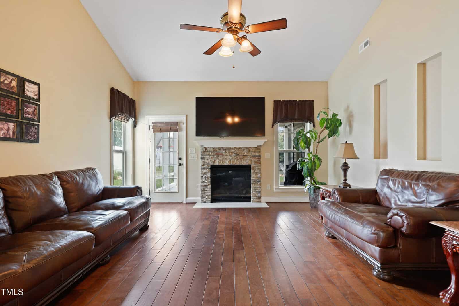 Living room with wood floors, two brown leather sofas, a stone fireplace, a mounted TV, ceiling fan, large windows, a potted plant, and a side table with a lamp. Walls are beige with three vertical niches on the right.