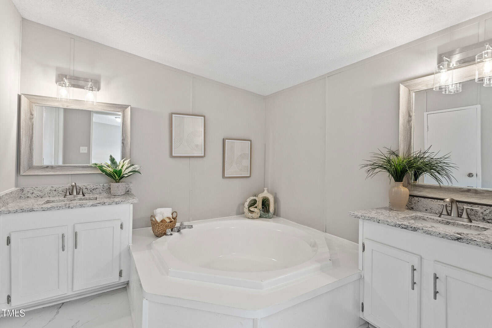 White bathroom with a large, central hexagonal bathtub surrounded by two vanities with granite countertops, mirrors, decorative plants, and framed wall art. The space has neutral walls and white cabinetry.