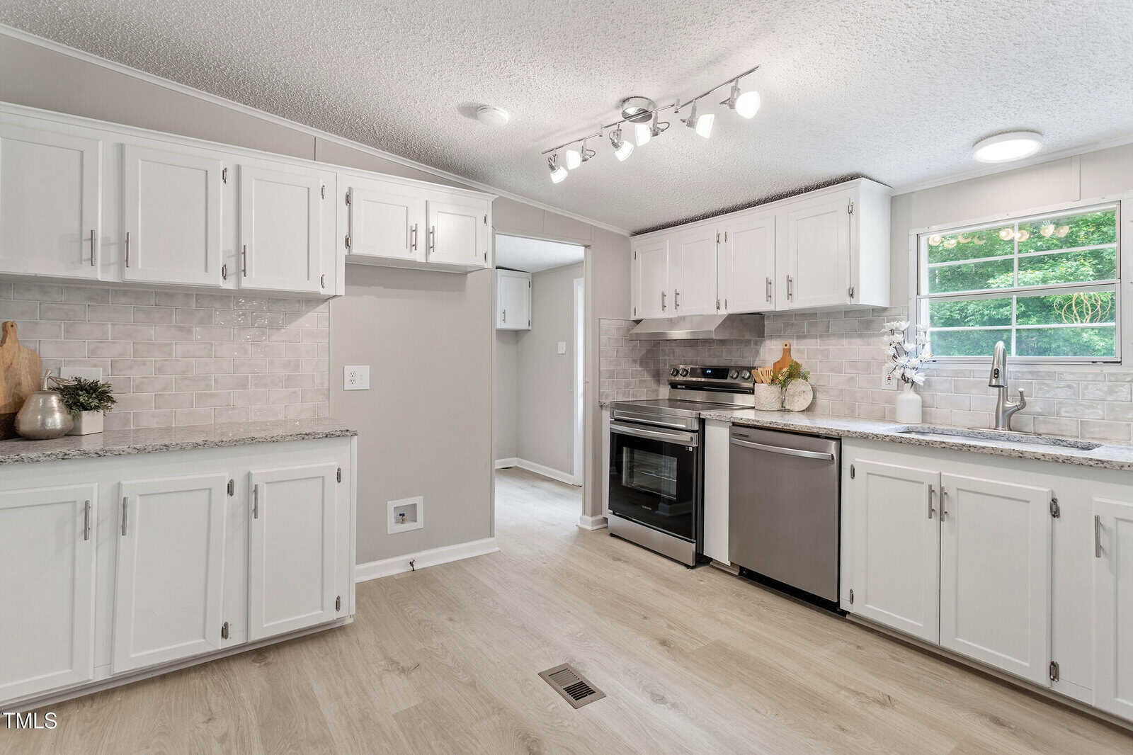 Modern kitchen with white cabinets, stainless steel appliances, light granite countertops, tile backsplash, and a large window above the sink letting in natural light. Light wood flooring throughout.