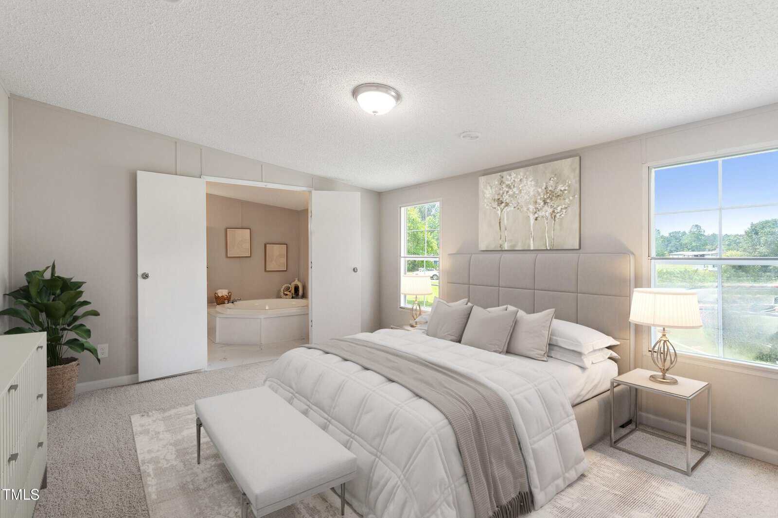 A bedroom with a large bed, nightstand with a lamp, a bench at the foot of the bed, a dresser with a plant, carpeted floor, and an open doorway leading to a bathroom with a bathtub and framed art.