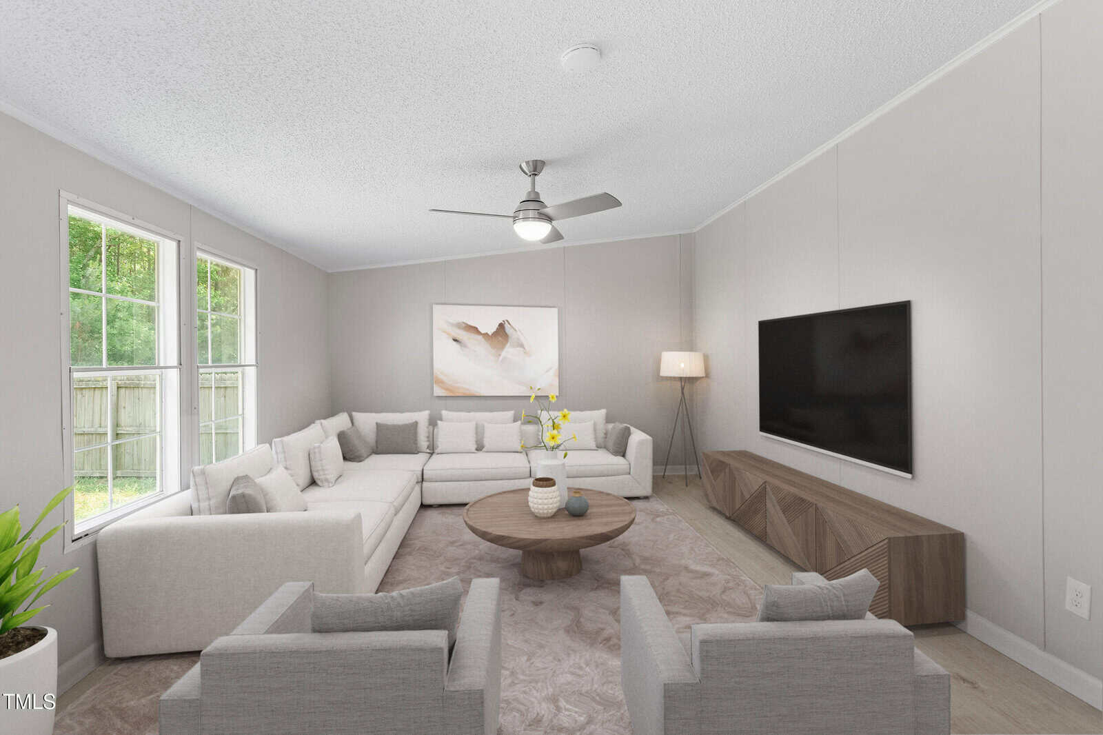 A modern living room with light gray walls, a large sectional sofa, two armchairs, a round coffee table, a TV on a wooden stand, a floor lamp, a wall art piece, and large windows letting in natural light.