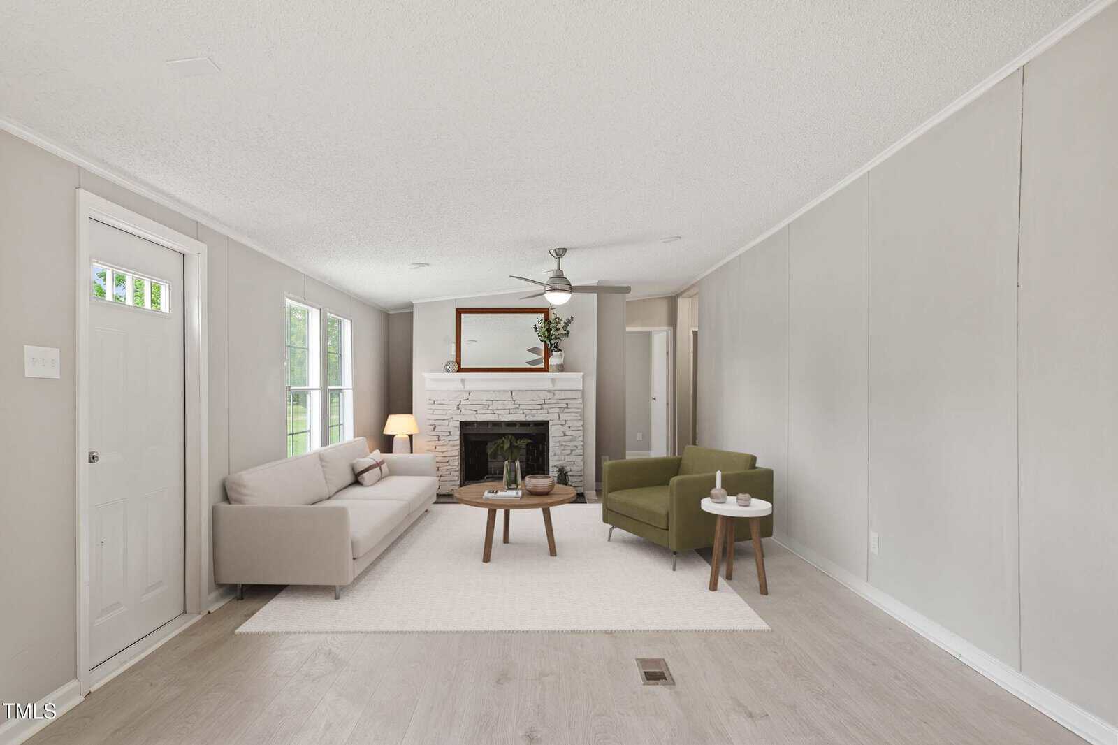 A modern living room with light gray walls, a beige sofa, green armchair, small coffee tables, white rug, and a fireplace with a mirror above it. Large windows and a door let in natural light.