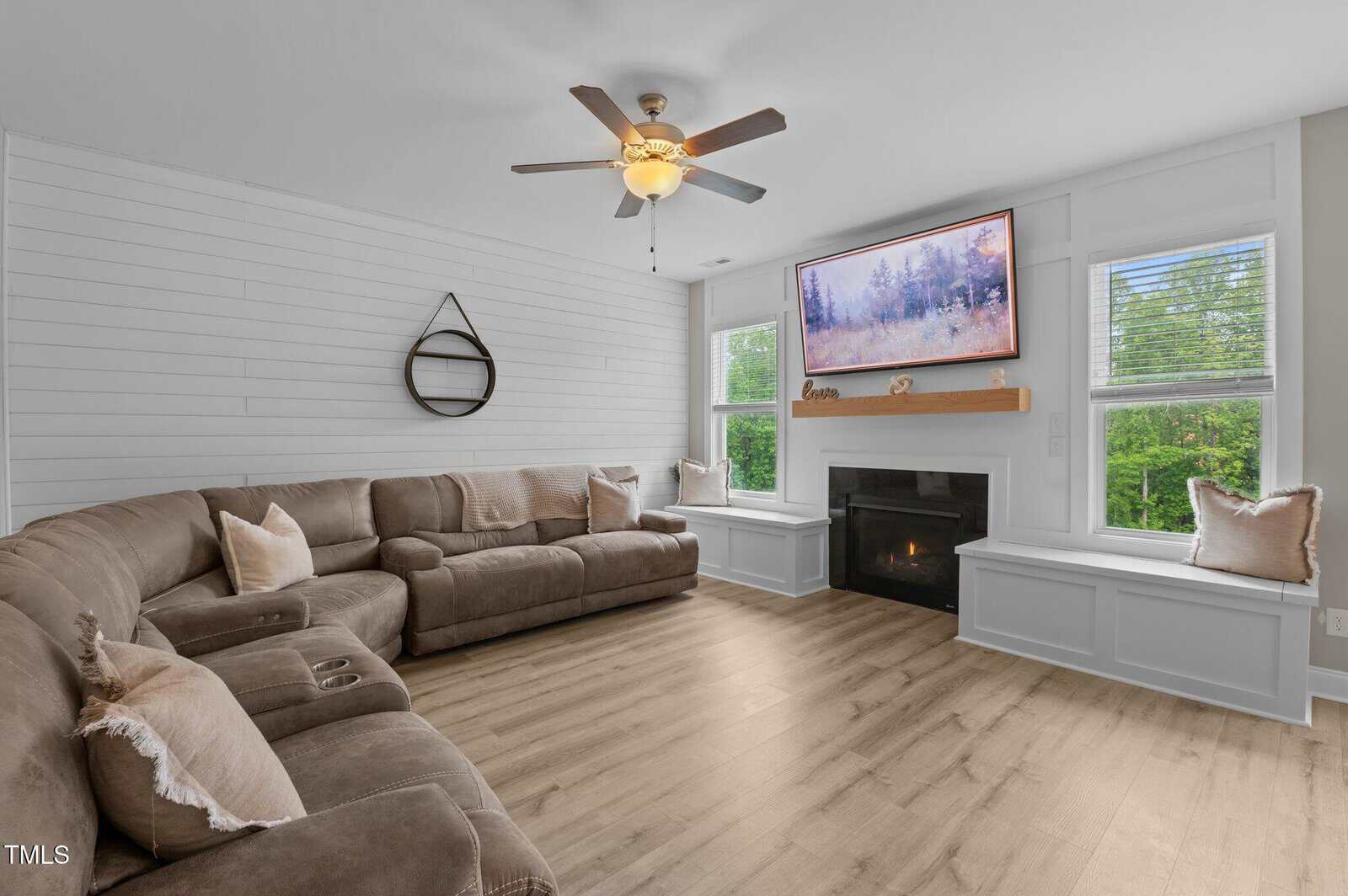 A living room with a brown sectional sofa, wood floors, a ceiling fan, a fireplace with a landscape painting above it, and two windows with window seats and pillows on either side of the fireplace.
