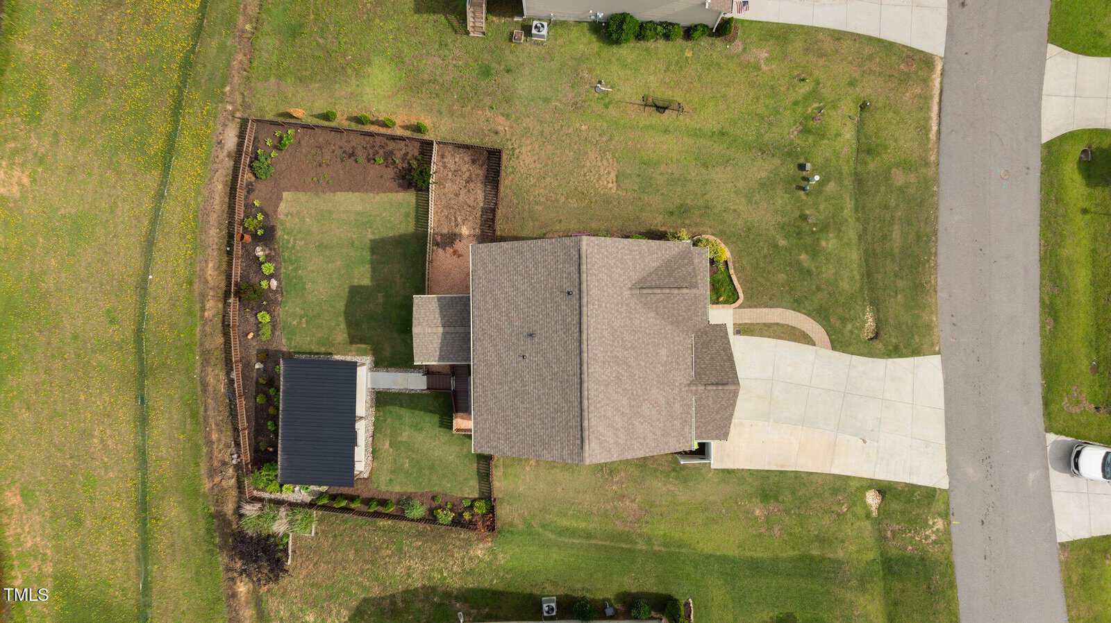 Aerial view of a suburban house with a neatly manicured lawn, a fenced backyard with garden beds, a driveway, and a car partially visible on the street. The house has a gray roof and is surrounded by green grass.