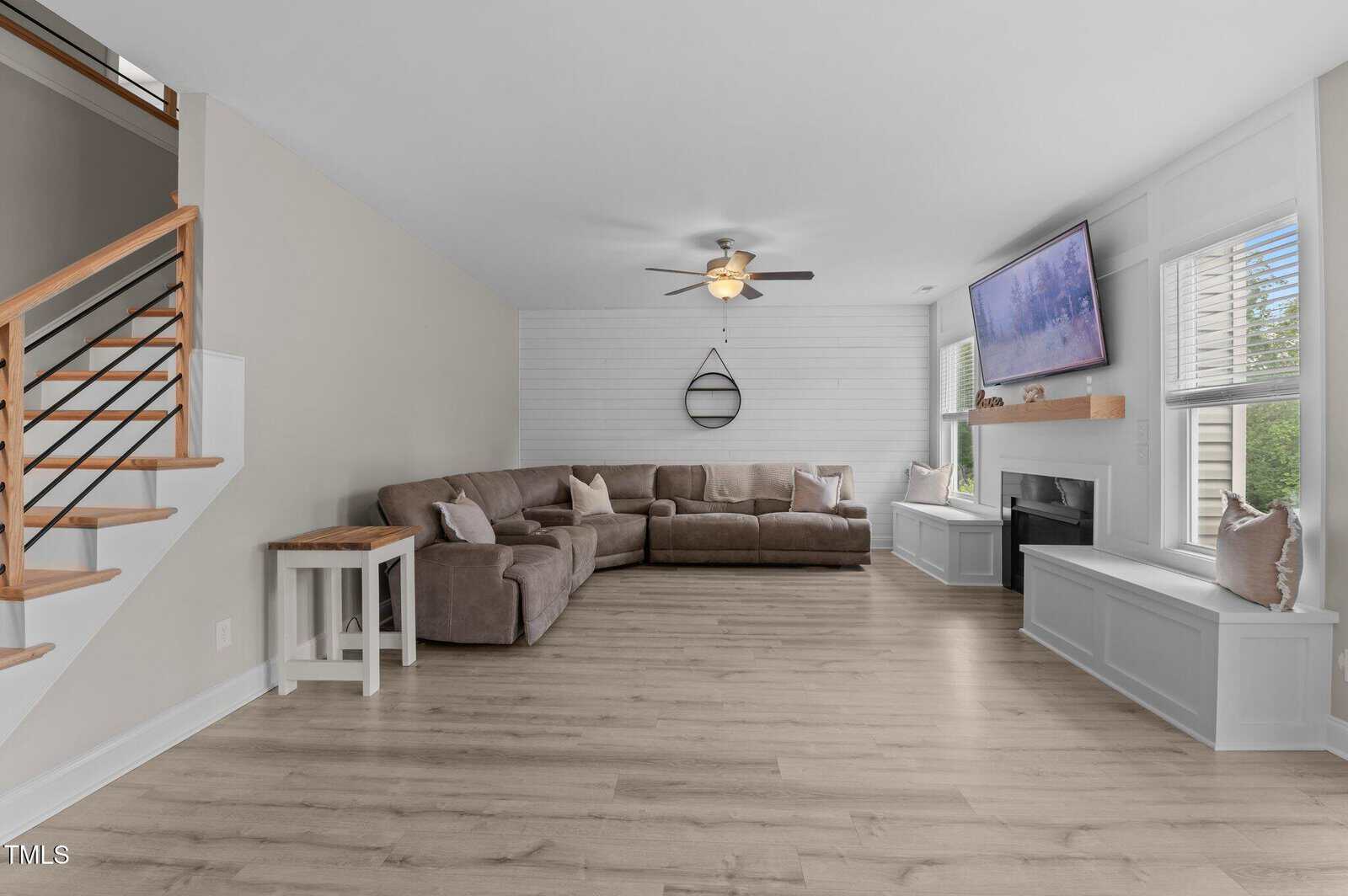 A spacious living room with light wood flooring, a large beige sectional sofa, accent pillows, a ceiling fan, a wall-mounted TV above a fireplace, built-in window seats, and a modern staircase with horizontal railings.