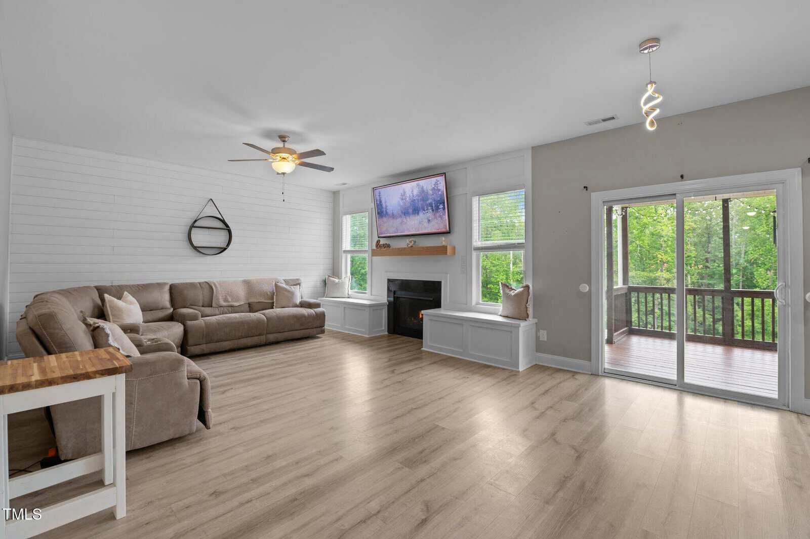 Modern living room with light wood flooring, a large beige sectional sofa, a wall-mounted TV above a fireplace, and sliding glass doors leading to a covered deck with views of green trees outside.