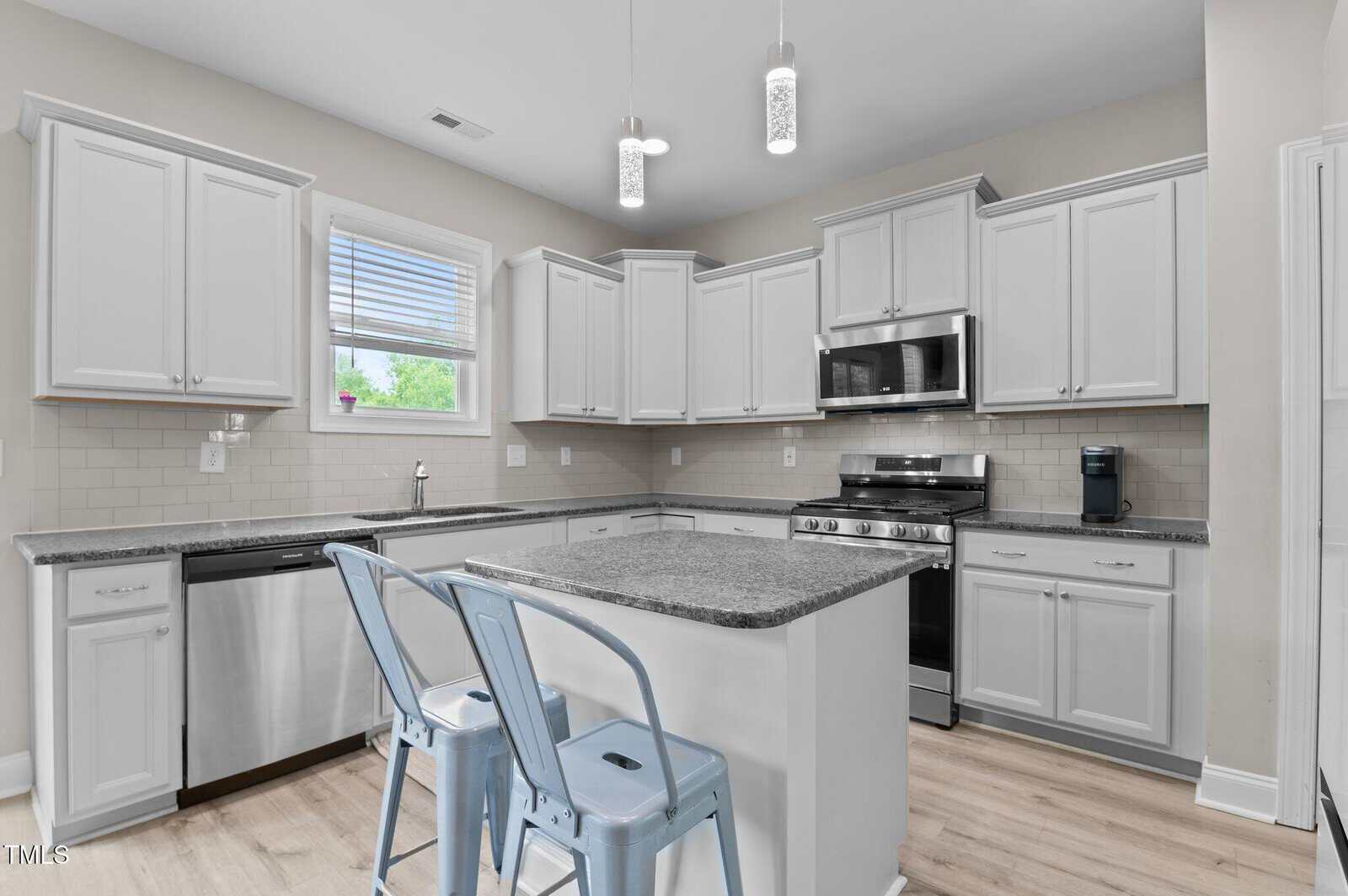 Modern kitchen with white cabinets, granite countertops, stainless steel appliances, light wood floor, and two blue metal stools at a central island. Pendant lights hang above the island, and a window is above the sink.