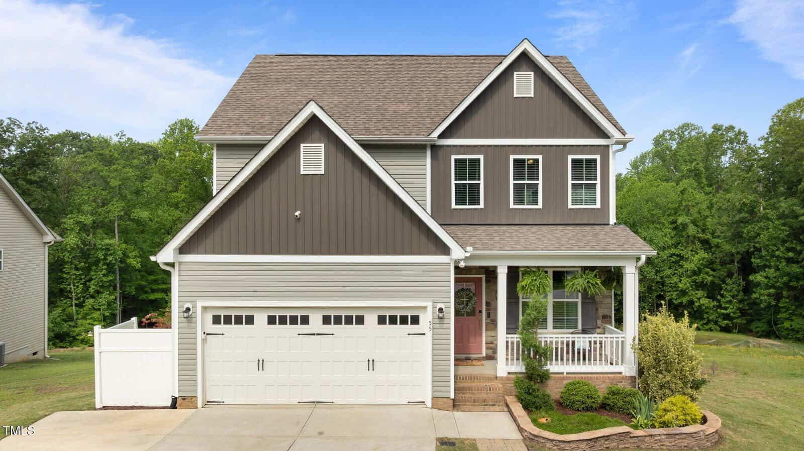 Two-story house with gray siding and dark brown accents, white garage door, front porch with white railings, potted plants, and a landscaped yard, set against a backdrop of green trees and a clear blue sky.