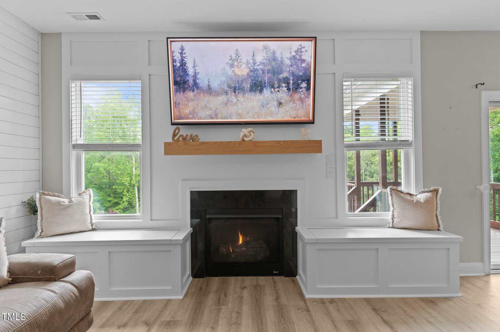 A cozy living room with a modern fireplace, a wooden mantel decorated with small items, and a landscape painting above. Built-in benches with cushions flank the fireplace, and large windows let in natural light.