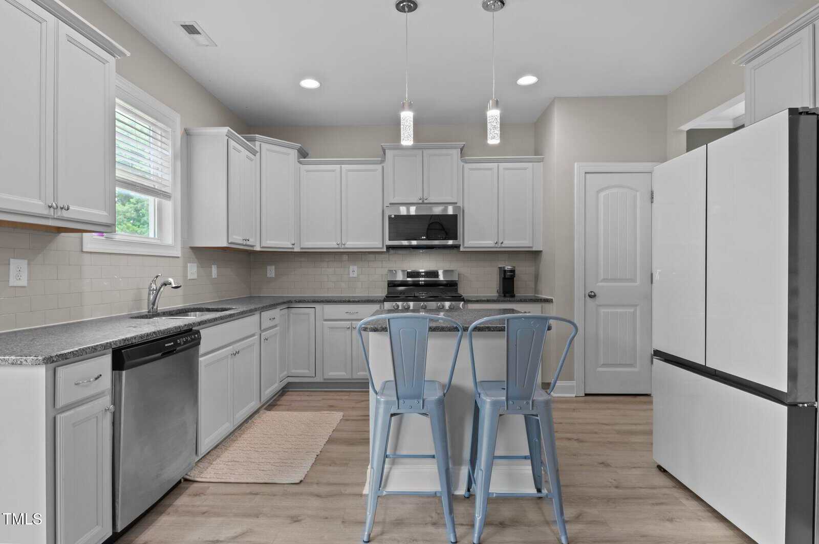 A modern kitchen with white cabinets, stainless steel appliances, a double-door fridge, a built-in microwave, two blue metal chairs at a gray breakfast bar, and pendant lights above the island.