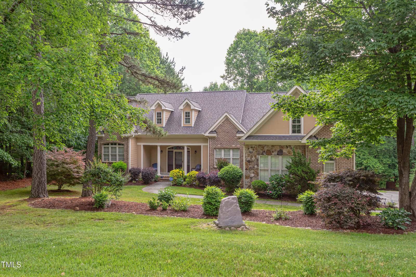 A two-story house with a stone and beige facade, gabled roof, and front porch, surrounded by manicured bushes, trees, and a green lawn with a large rock in the foreground.