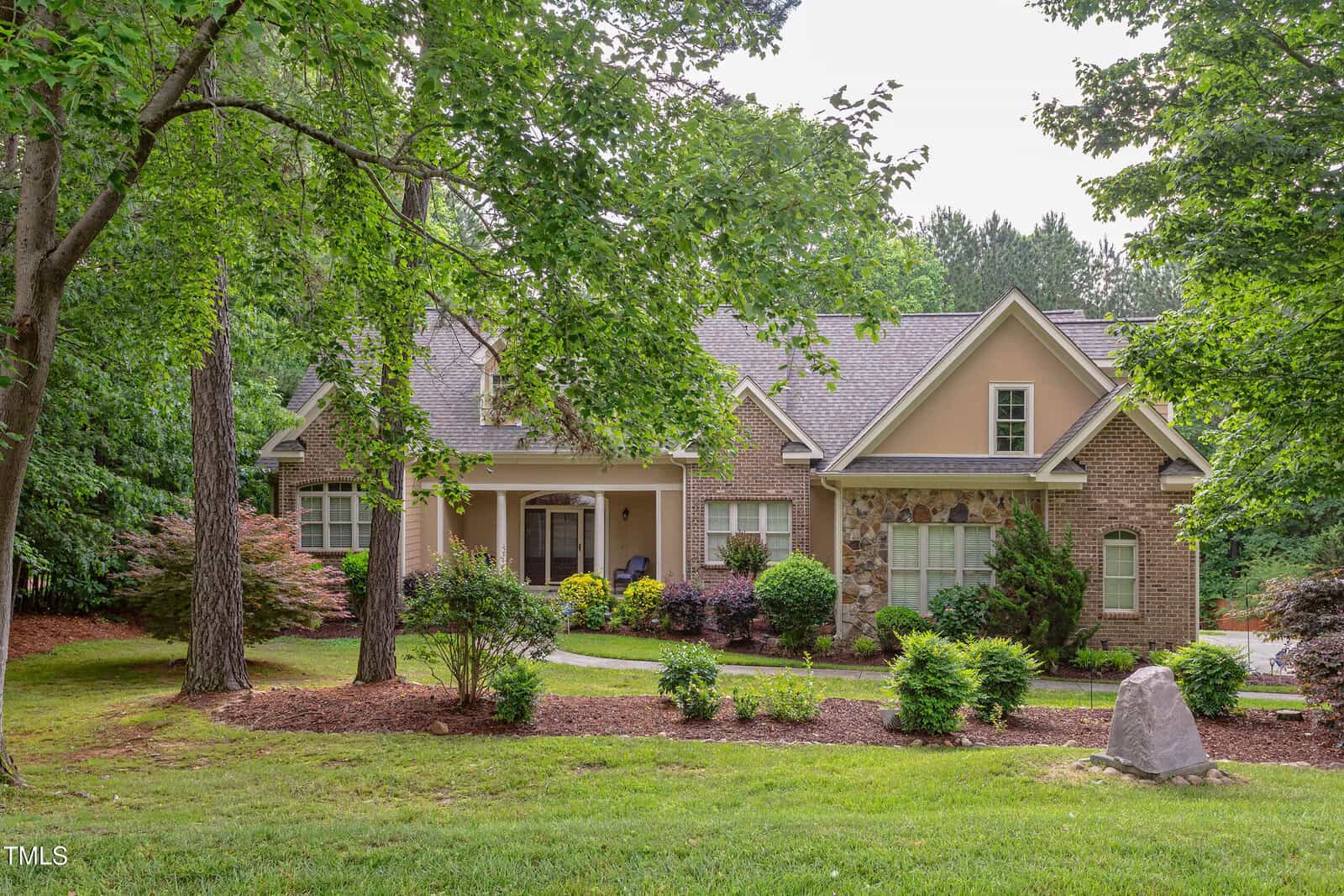 A two-story suburban house with tan siding and brick accents sits at 504 Belmont Circle, Wake Forest. Surrounded by trees and landscaped shrubs, the home features a grassy yard with a stone near the front and a partially visible driveway on the right.