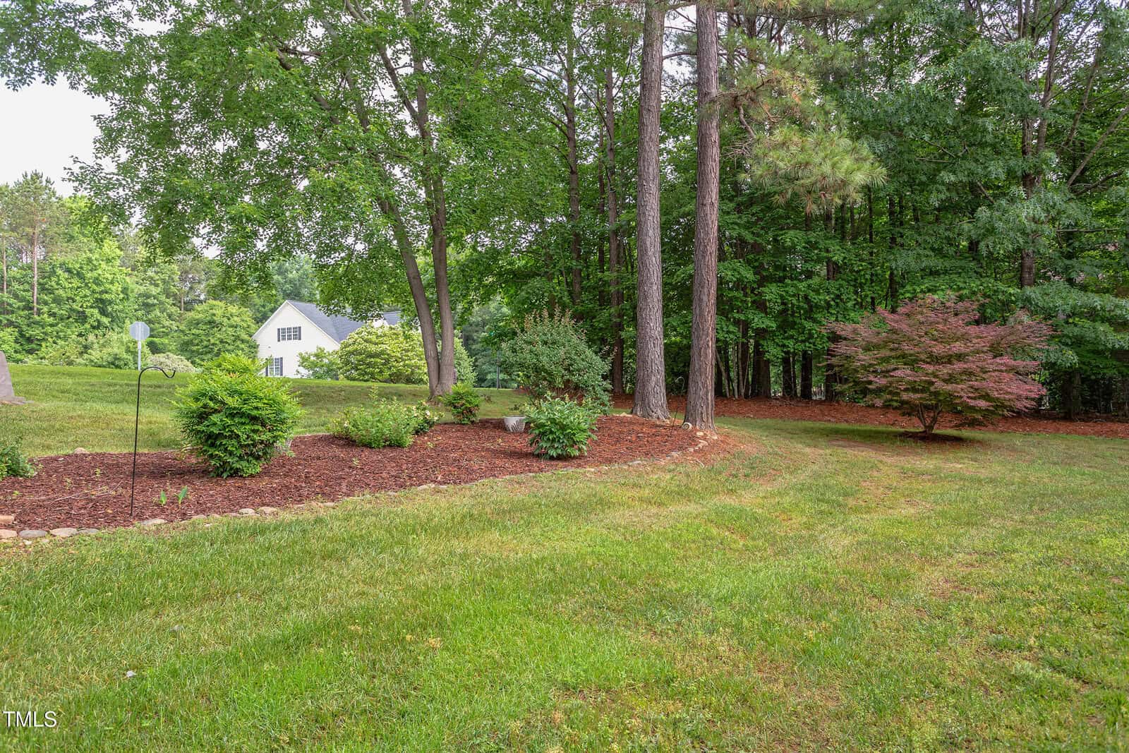 A landscaped yard with green grass, mulch, shrubs, and several mature trees. In the background, a white house is partially visible among trees, and the area appears well-maintained and bordered by woods.