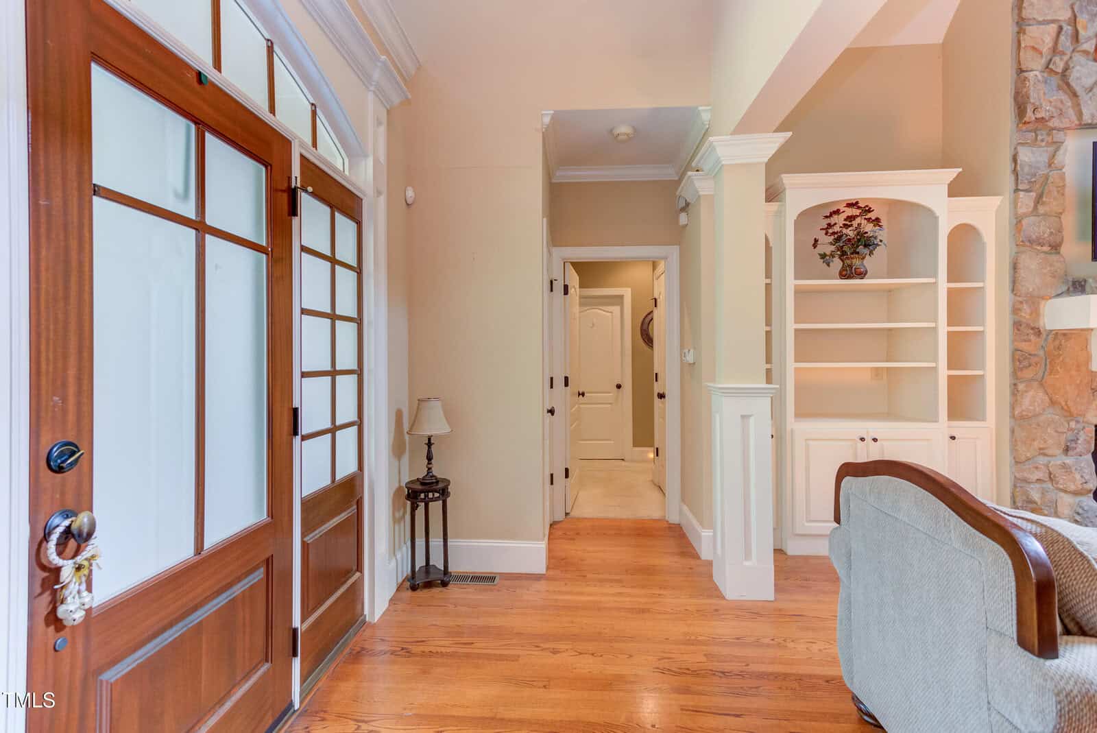 A home entryway with a wooden front door and glass panels, hardwood floors, beige walls, a small table with a lamp, built-in shelves, and a partial view of a stone fireplace and sofa.