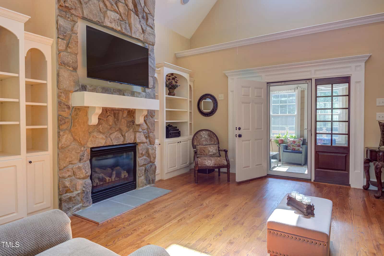 A living room with a stone fireplace, mounted TV, built-in white shelves, wooden floors, armchair, ottoman, and open doors leading to a sunroom with plants and large windows letting in sunlight.