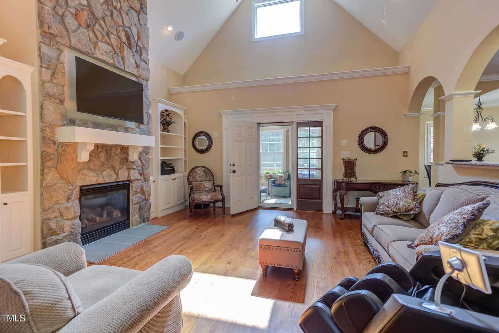 A living room with a stone fireplace and mounted TV, built-in shelves, wood floors, large windows, a sofa, armchair, and massage chair. French doors lead to a sunroom; the kitchen is visible through an arched opening.