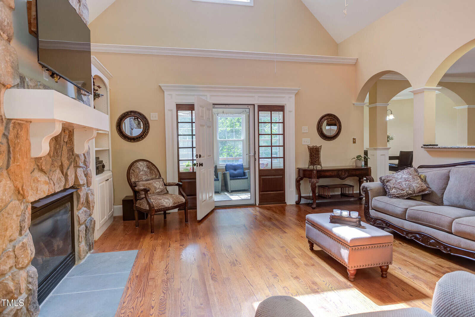 A living room with wood floors, a stone fireplace, and neutral-colored furniture. French doors open to a sunroom with large windows. The space is bright with natural light and decorated with classic chairs and wall mirrors.