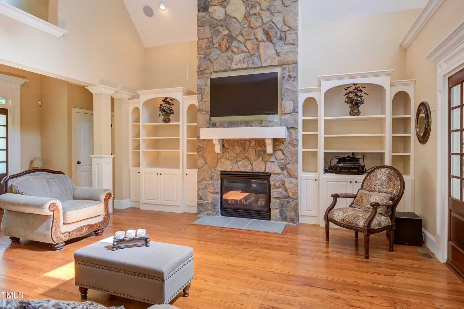 A living room with hardwood floors, a stone fireplace, a TV mounted above the mantel, built-in white shelves and cabinets, and two upholstered armchairs. Natural light brightens the space.