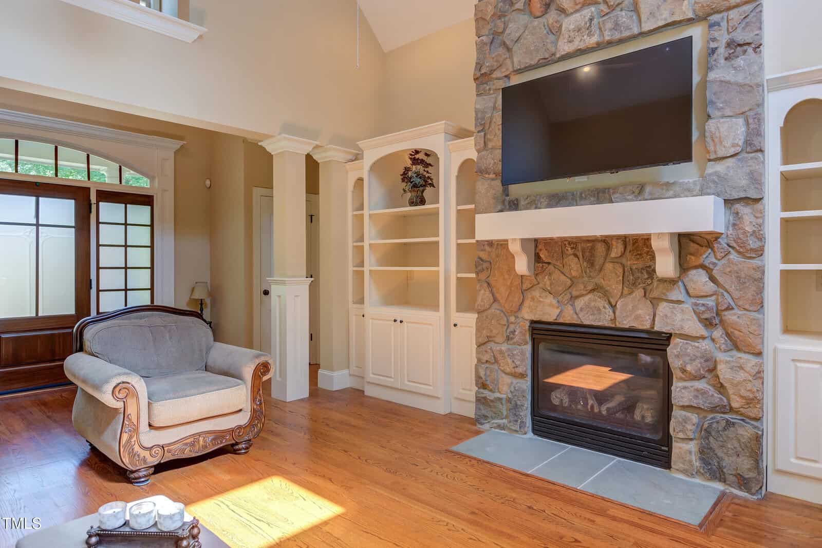 A living room with a stone fireplace, a mounted flat-screen TV, built-in white shelves, a gray upholstered armchair, hardwood floors, and large windows near the entrance door.