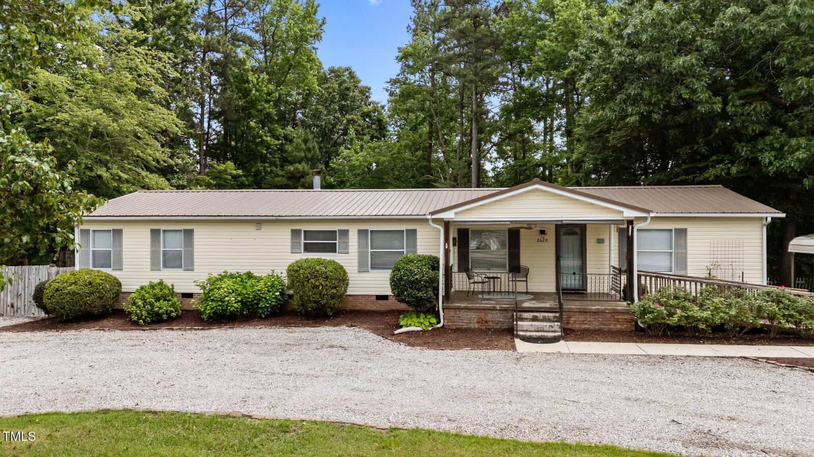 Charming single-story beige manufactured home at 2620 Hicksboro Road, Henderson, featuring a metal roof, front porch with ramp and steps, surrounded by shrubs and trees, and a gravel driveway in front.