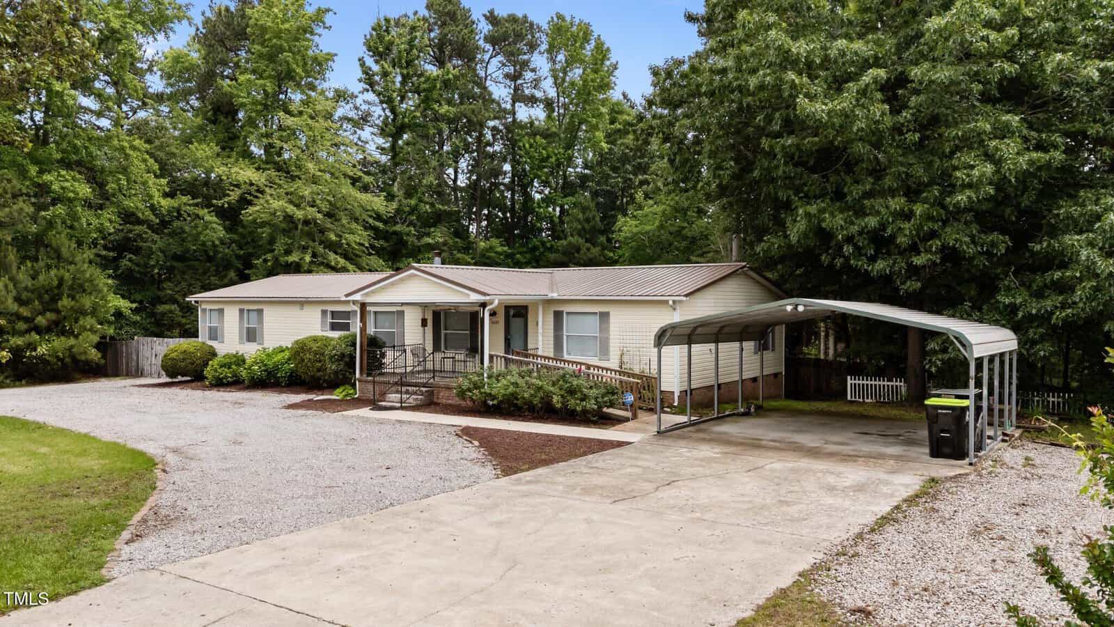 Single-story beige house with a metal roof, front porch with railing, gravel driveway, and attached carport on the right. The yard is bordered by trees and bushes, with a wooded area in the background.