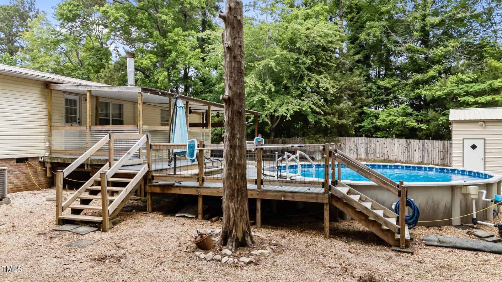 Backyard with a raised wooden deck featuring two staircases, an above-ground pool with railing, a tree in front, gravel ground cover, and a fenced yard with trees and a shed in the background.
