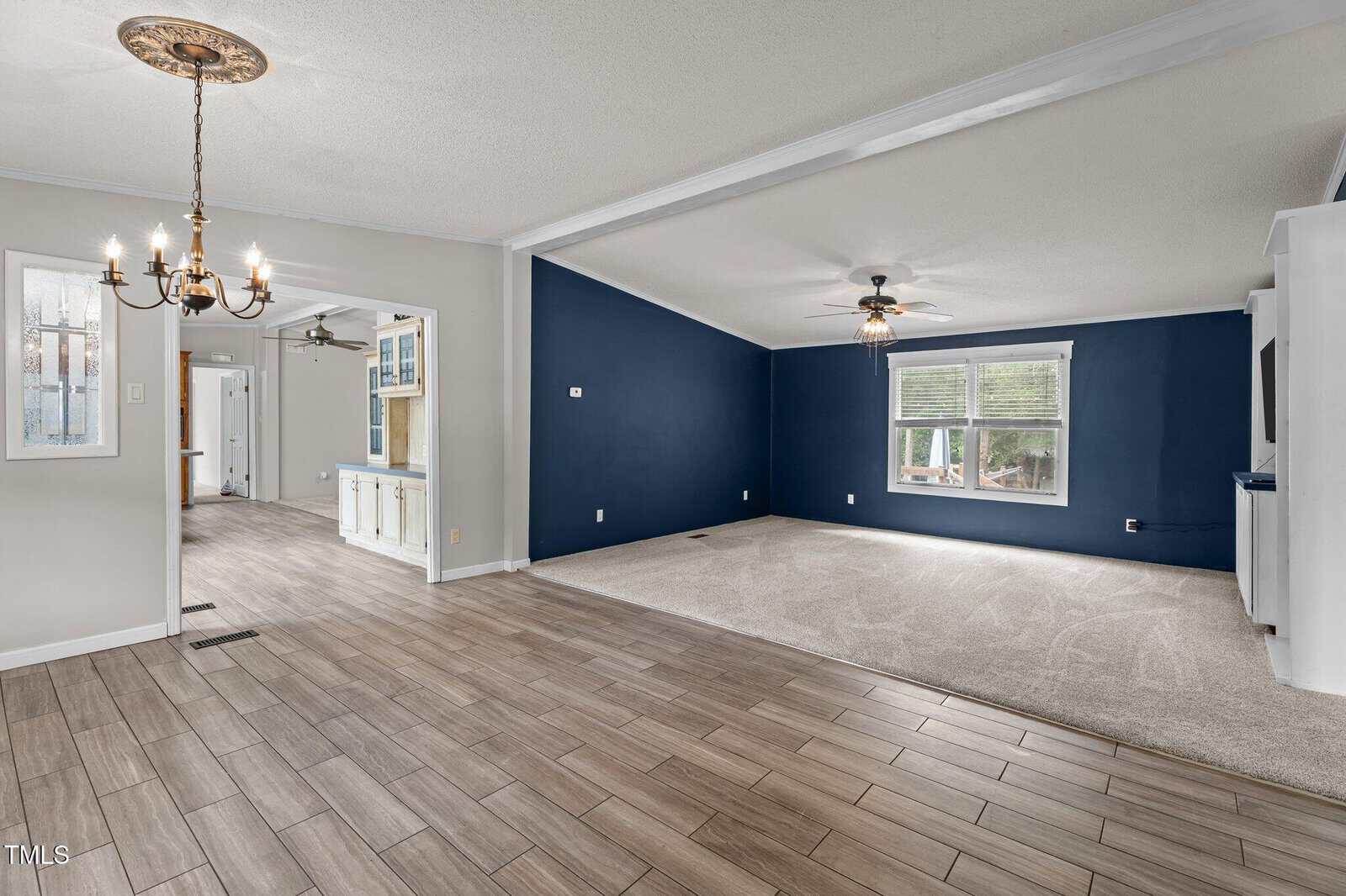 Open living area with tile and carpet flooring, navy accent wall, ceiling fans, white trim, and a chandelier. A dining area leads to a bright hallway and kitchen with glass-front cabinets in the background.