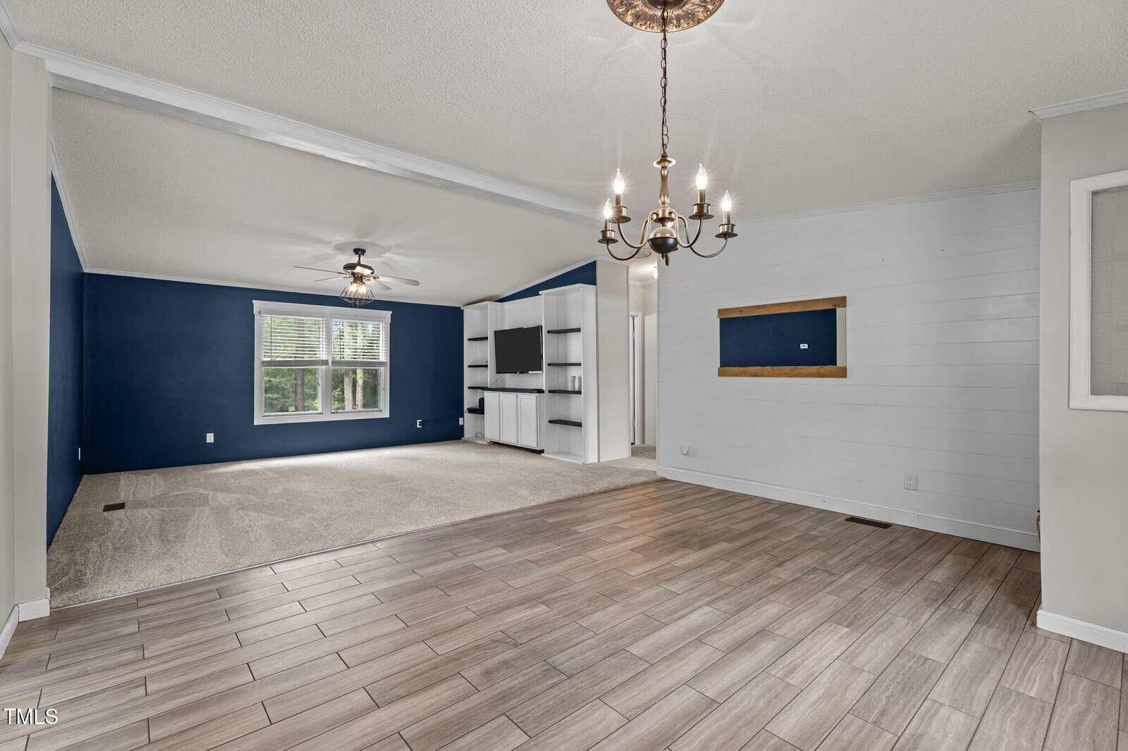 A living area with a tiled floor transitions into a carpeted section. The room has blue and white walls, a ceiling fan, a chandelier, large window, built-in shelving, and a wooden-framed niche on the wall.