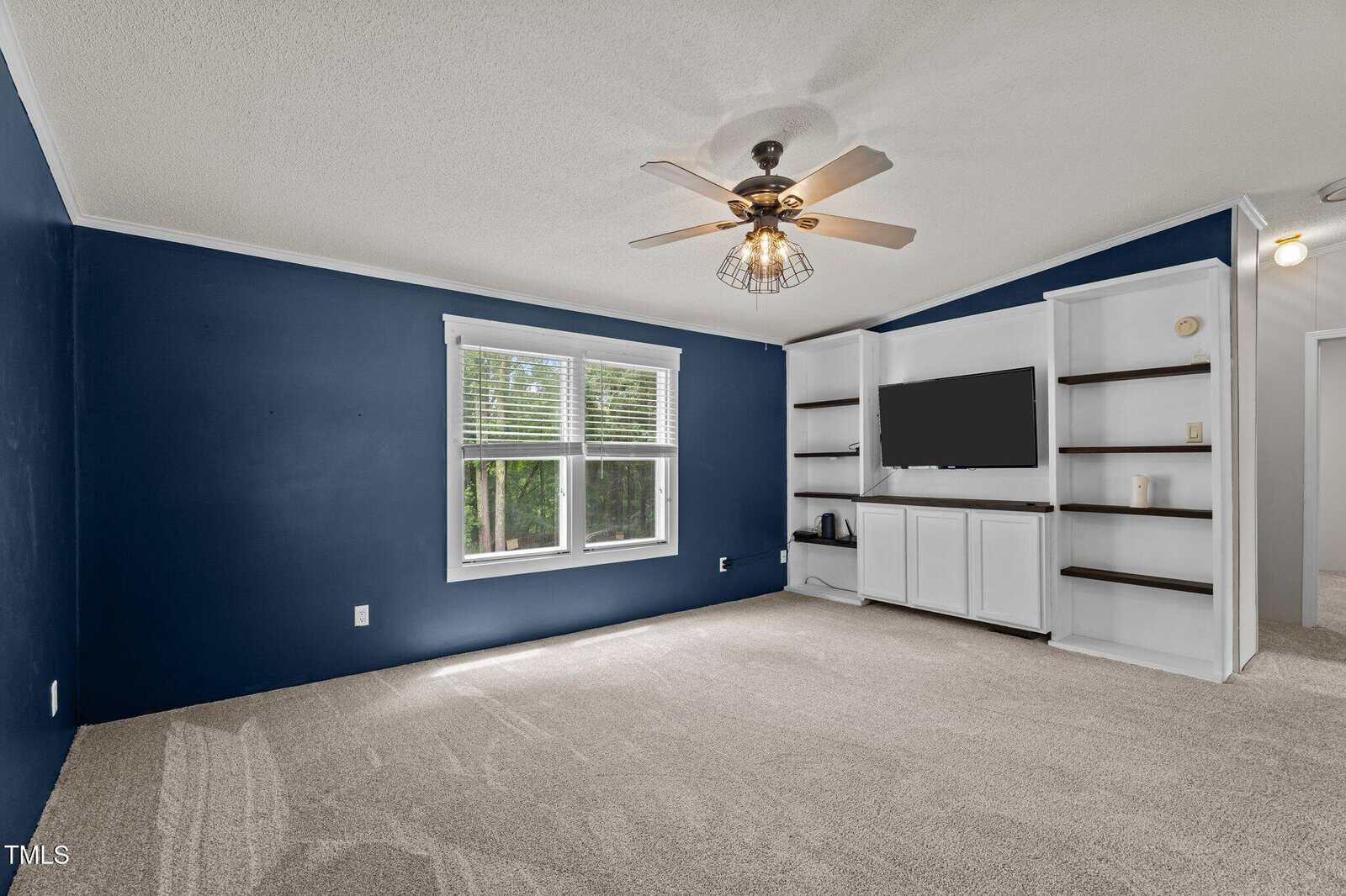 A room with blue accent walls, beige carpet, a ceiling fan with a light, a large window, and built-in white shelving and cabinets with a mounted TV. The room is empty of furniture.