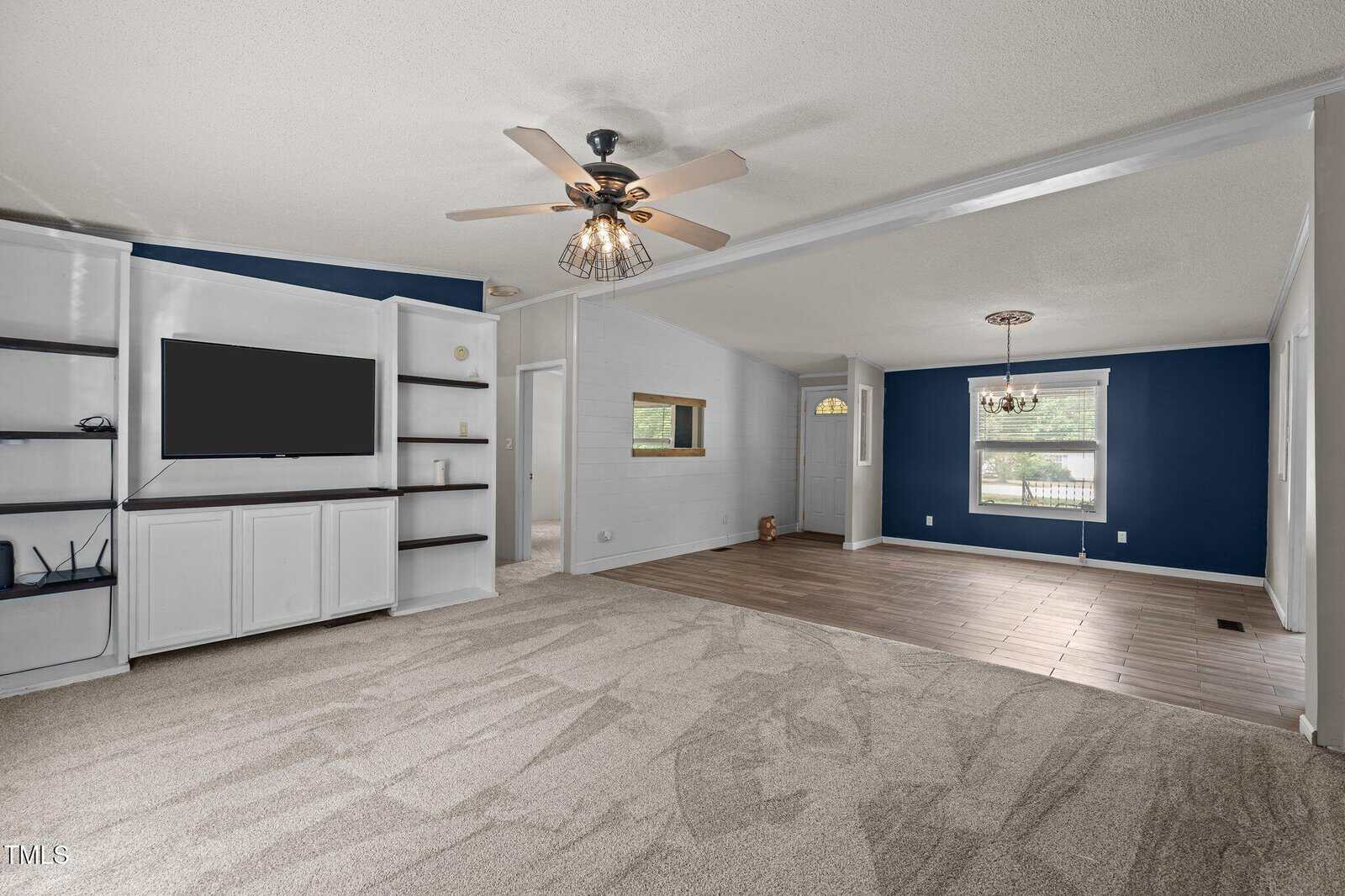 A spacious living room with built-in white shelves and a TV on the left, beige carpet, a ceiling fan, and an adjoining dining area with blue accent walls, a window, and tile flooring.