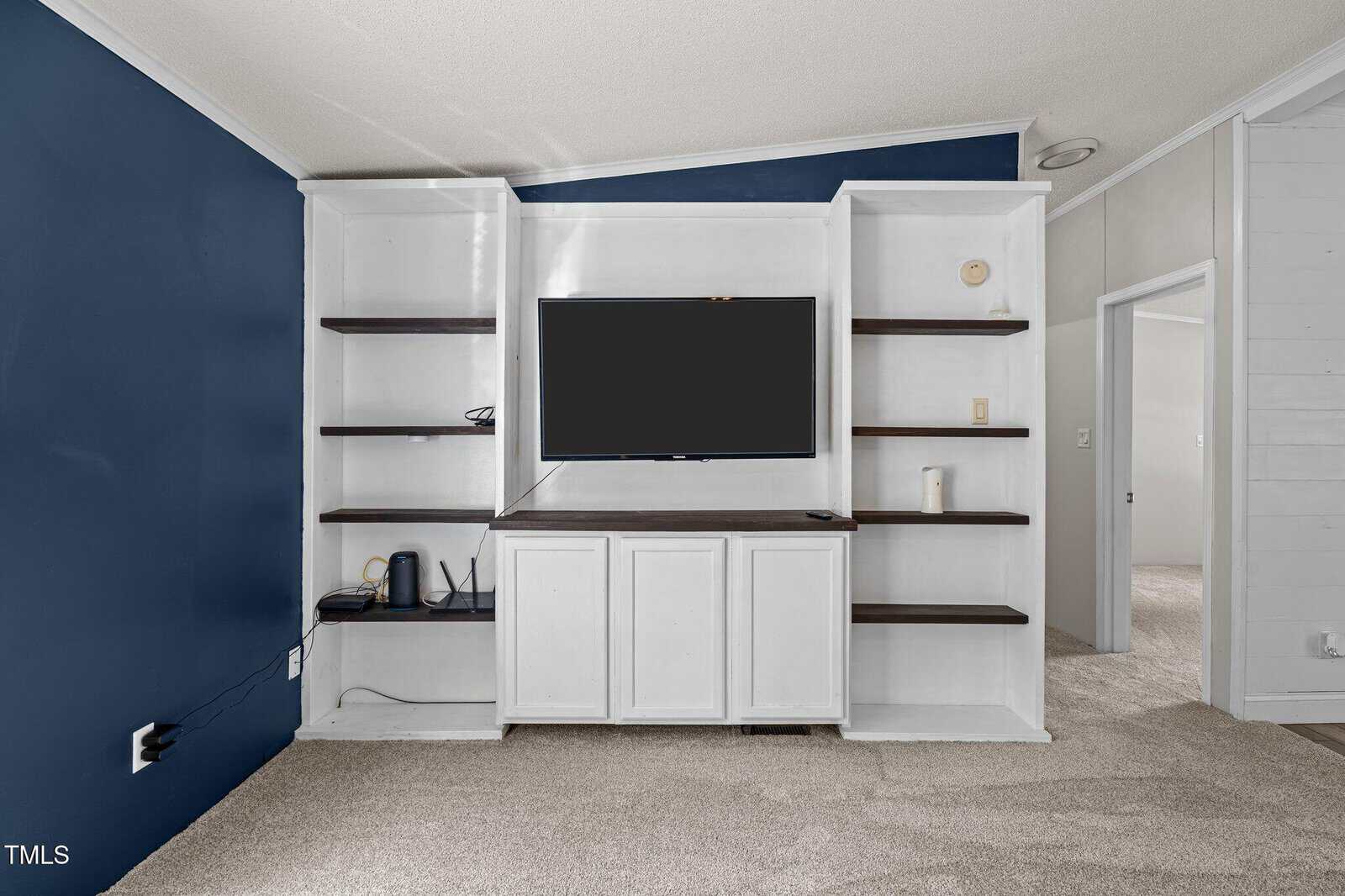 A built-in white entertainment center with dark wood shelves holds a flat-screen TV, a modem, a router, and a small speaker. The walls are painted navy blue and white, and the floor is carpeted in beige.