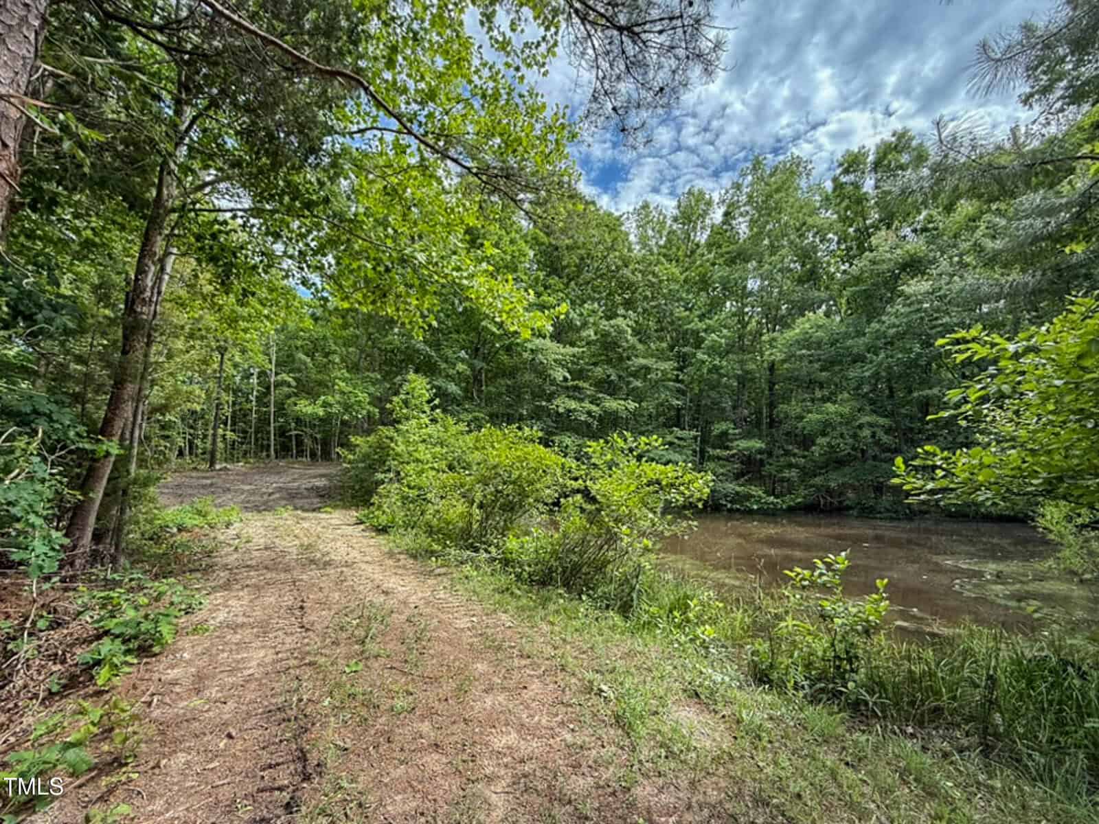A dirt path runs alongside a small pond near 9016 Aaron Creek Church Road in Oxford, surrounded by dense green trees under a partly cloudy sky. Vegetation is lush, giving the area a natural and untouched feel.