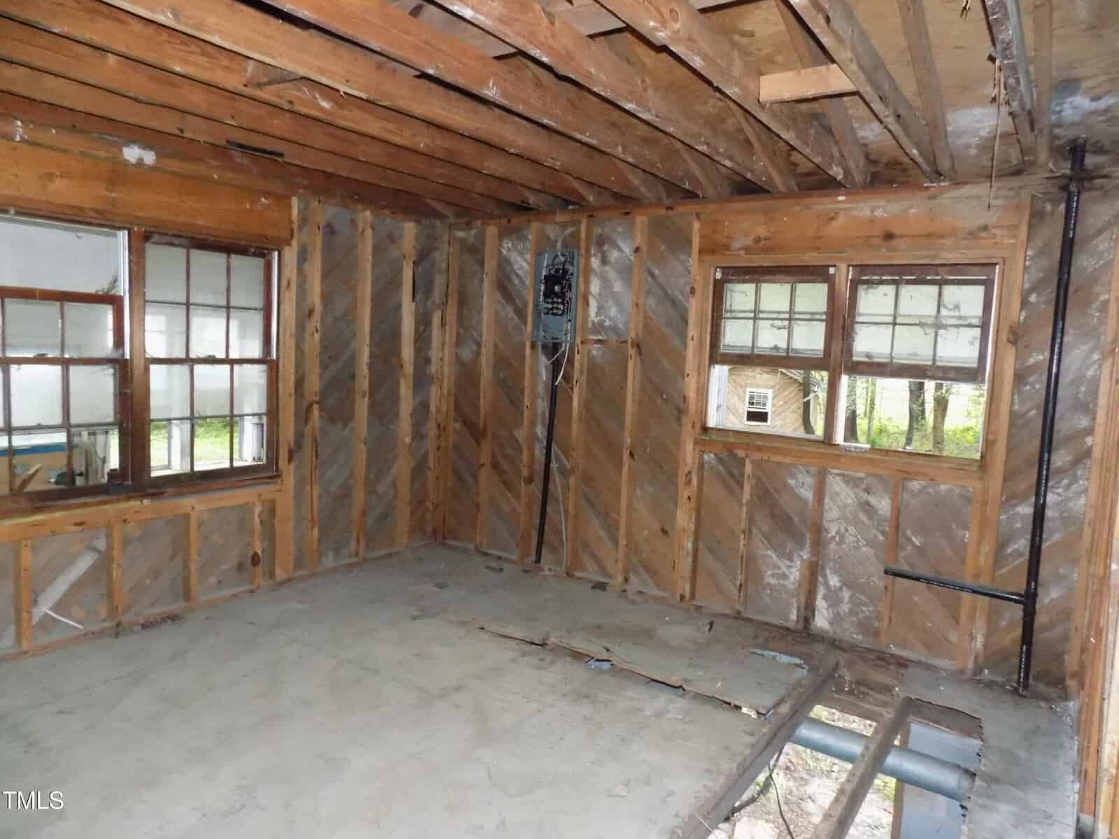 An interior view of a room under construction, featuring exposed wooden wall studs, ceiling joists, and a concrete floor with a hole exposing subfloor components. Two windows are present, and an electrical breaker box is mounted on the wall.
