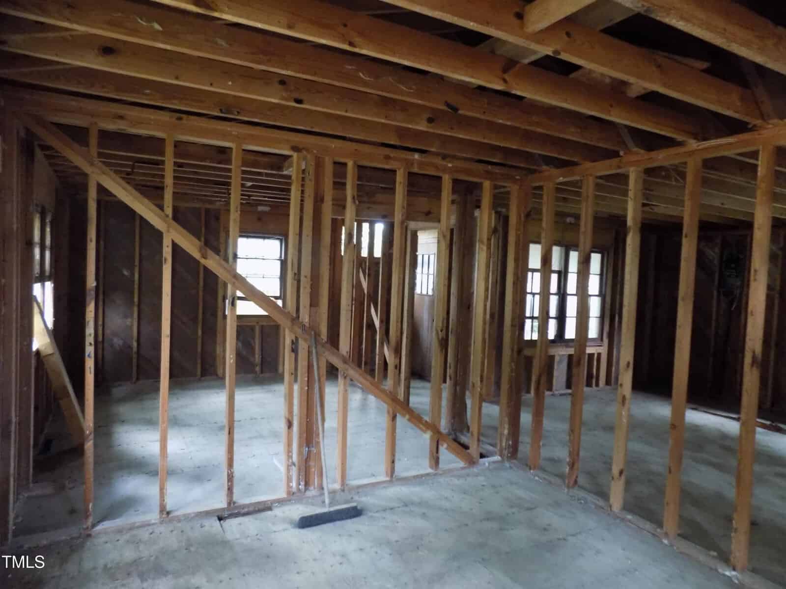 Exposed wooden wall studs and ceiling beams in a house under renovation, with unfinished floors and visible windows in the background. No drywall or insulation is present.