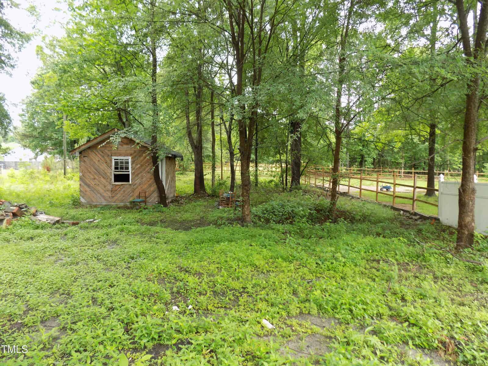 A small wooden shed with a single window sits among tall trees and green grass in a fenced backyard, with scattered debris and a white fence visible in the background.
