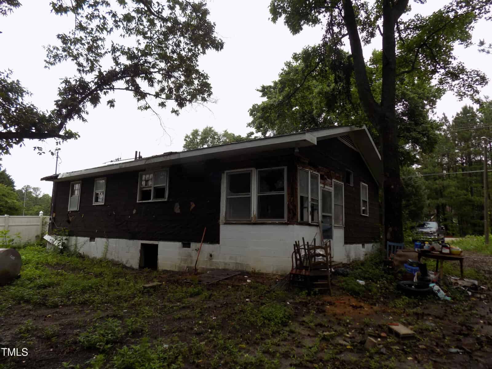 A weathered, single-story house with dark siding, damaged windows, and a white cinder block foundation sits surrounded by overgrown grass and trees on a cloudy day. Worn furniture and debris are scattered near the entrance.