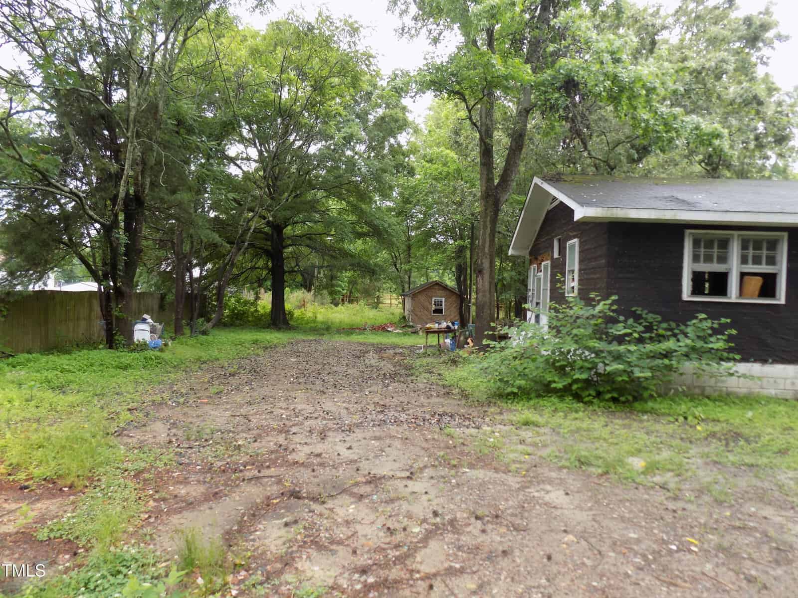 A dirt driveway leads to a small brown house with white trim surrounded by green trees and bushes; a wooden shed is visible in the background.
