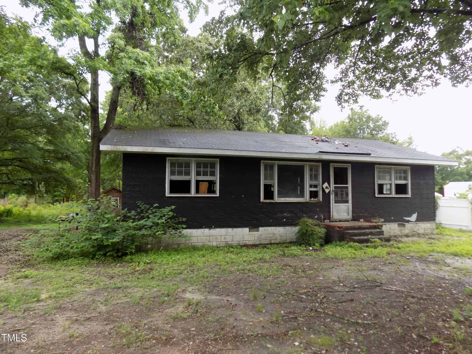 A small, single-story black house with white trim sits in a yard with overgrown grass and trees. The roof has visible damage, and the front steps lead up to a door with a geometric window.