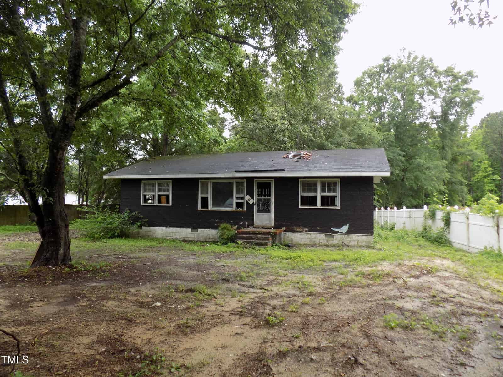 A small, black-painted house with a damaged roof sits in a yard with patchy grass and dirt. Large trees surround the house, and a white fence runs along the right side. The scene appears overcast and damp.