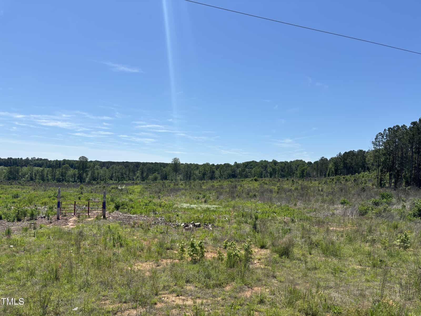 A wide, open grassy field with sparse vegetation and a small fenced area on the left. Trees line the horizon under a clear blue sky with a thin, bright sunbeam visible. A power line crosses the upper right corner.