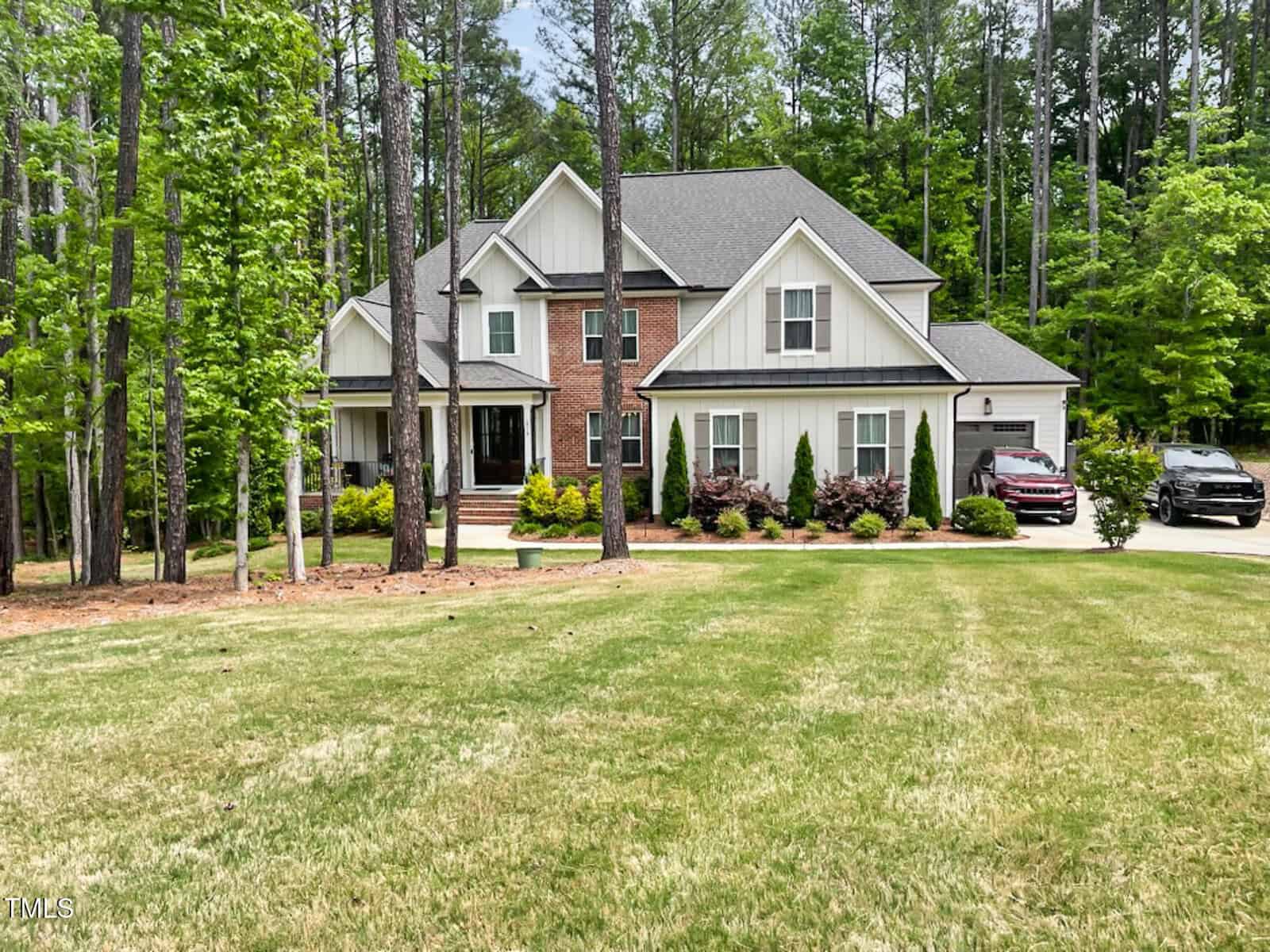 A two-story suburban house with brick and white siding at 1218 Rogers Farm Road, Wake Forest, is surrounded by tall trees. The front yard features trimmed grass and shrubs, with two cars parked in the driveway on the right side.