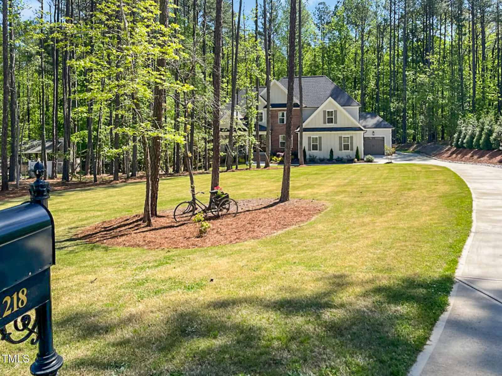 A two-story house with a front porch sits at the end of a curved driveway surrounded by trees and grass. A black mailbox numbered 218 is in the foreground, and a bicycle leans against a tree in the yard.